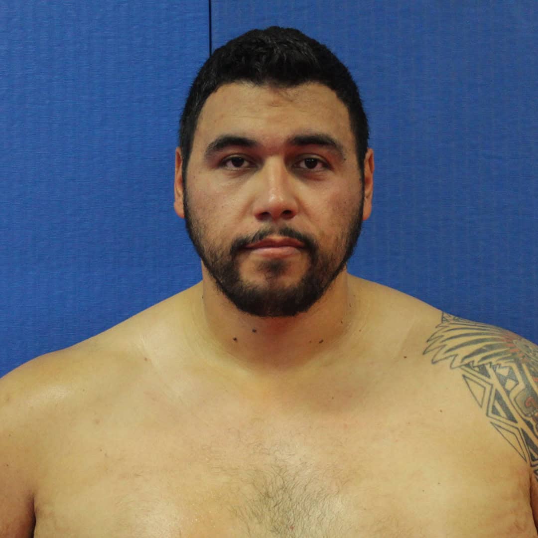 Sumo wrestler with short black hair and beard, blue background.