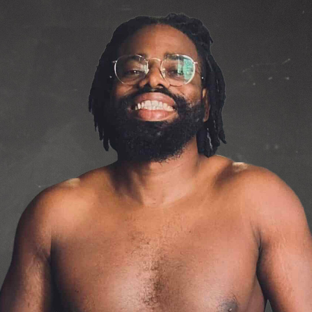 Smiling man with glasses and dreadlocks, shirtless, against dark background.